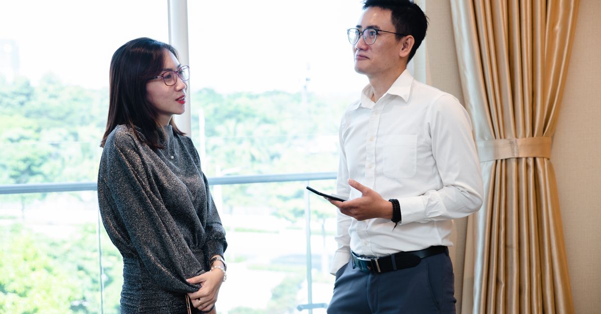 Two colleagues engaged in a business conversation indoors with a window view.