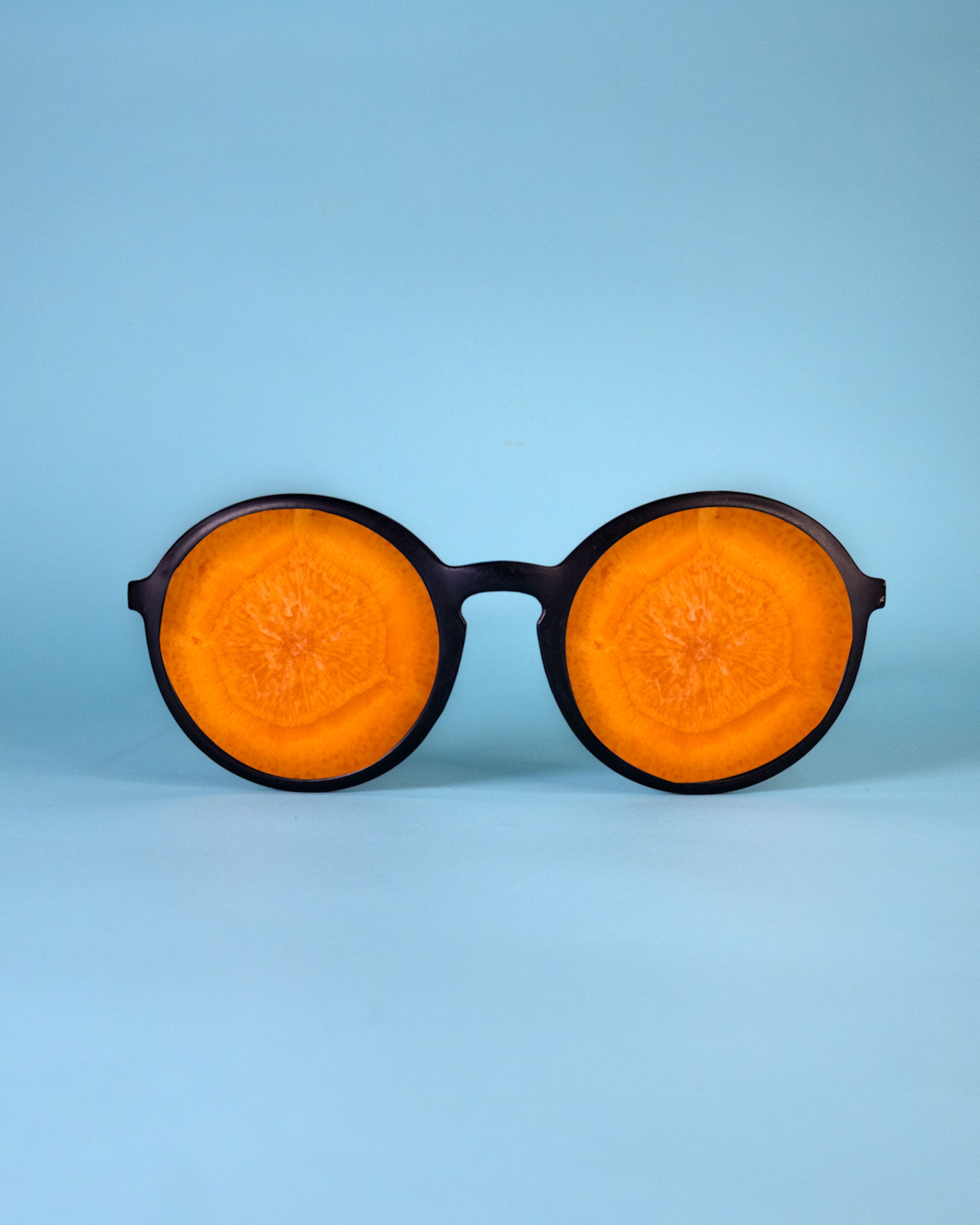 Artistic composition of eyeglasses with carrot slices as lenses against a blue backdrop.