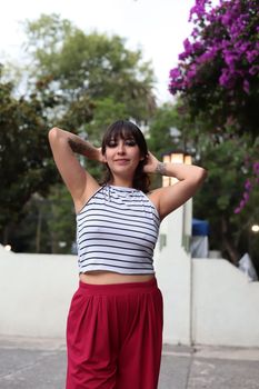 Smiling young woman posing outdoors in casual attire, surrounded by vibrant flowers and greenery.