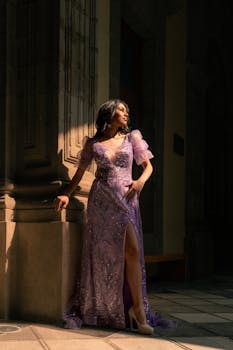 Woman in a beautiful purple dress poses elegantly in a sunlit architectural setting.