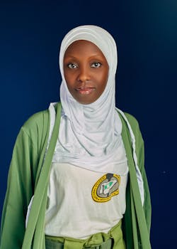 Portrait of a young woman wearing a white hijab and traditional green attire against a blue background.