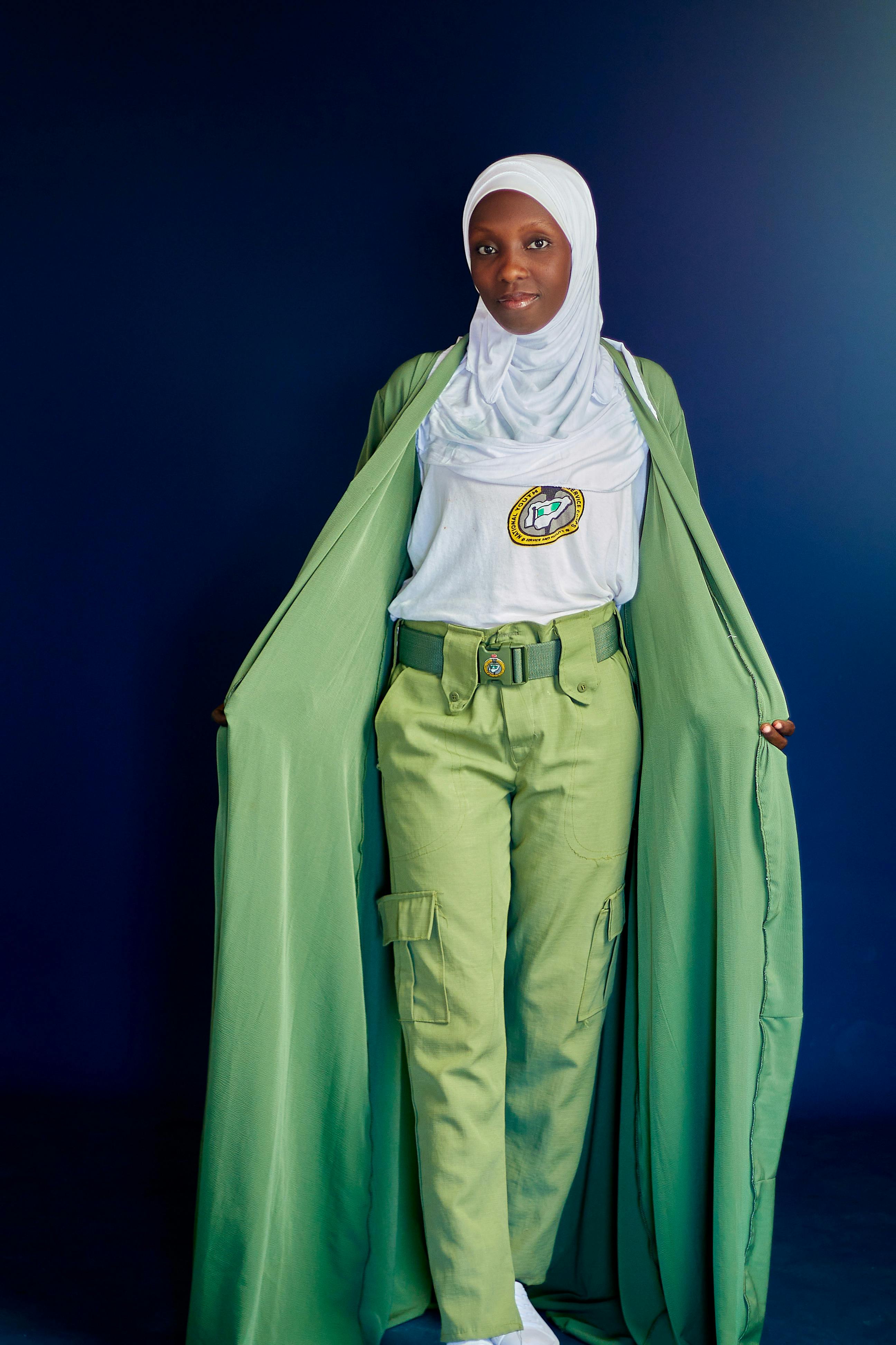 Confident young woman in green cape and traditional hijab posing against blue backdrop.
