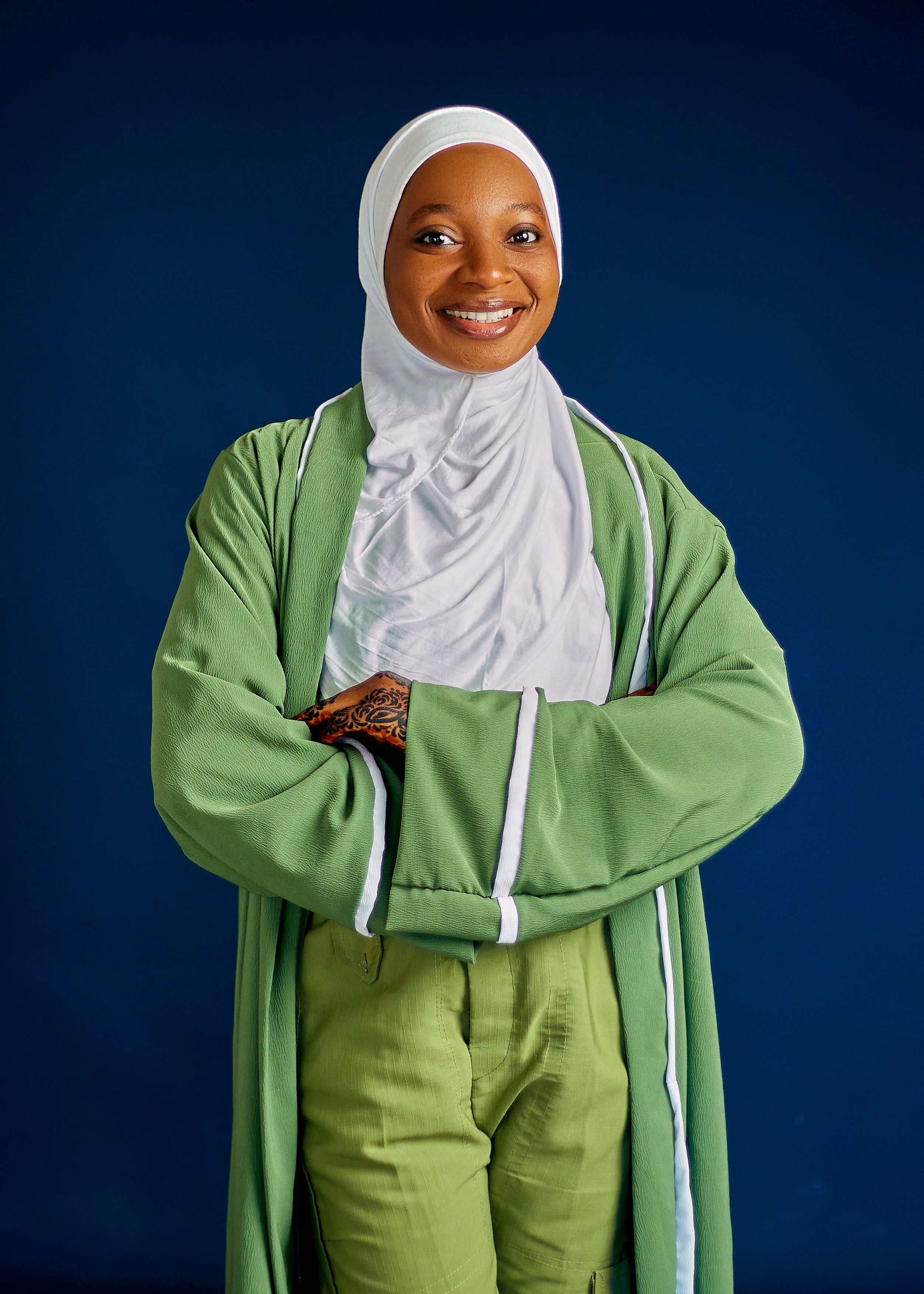 Confident woman wearing a green outfit and white hijab, smiling with arms crossed.