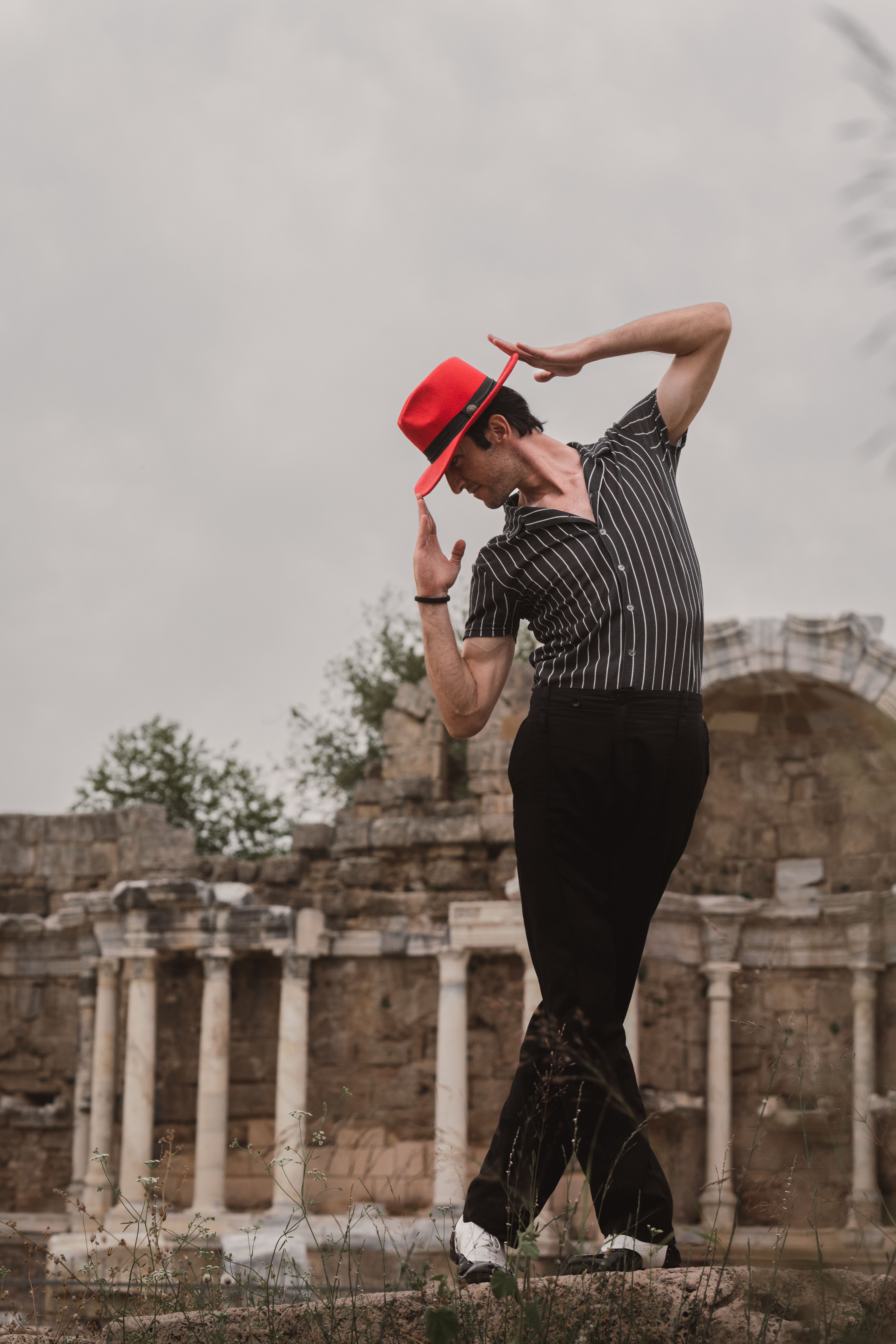 Stylish dancer striking a dramatic pose in front of ancient ruins, wearing a red hat and striped shirt.
