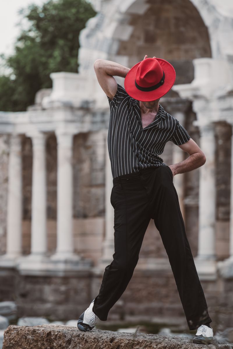 Man elegantly posing with red hat and striped shirt in front of ancient European architecture.