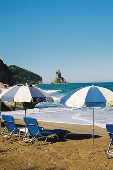 A picturesque beach with blue umbrellas, chairs, and rocky outcrops on a sunny day with clear skies.