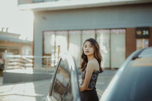 Stylish young woman standing by a car in an urban area with sunlight.
