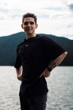 Casual portrait of a young man in front of a serene mountain lake with cloudy sky.