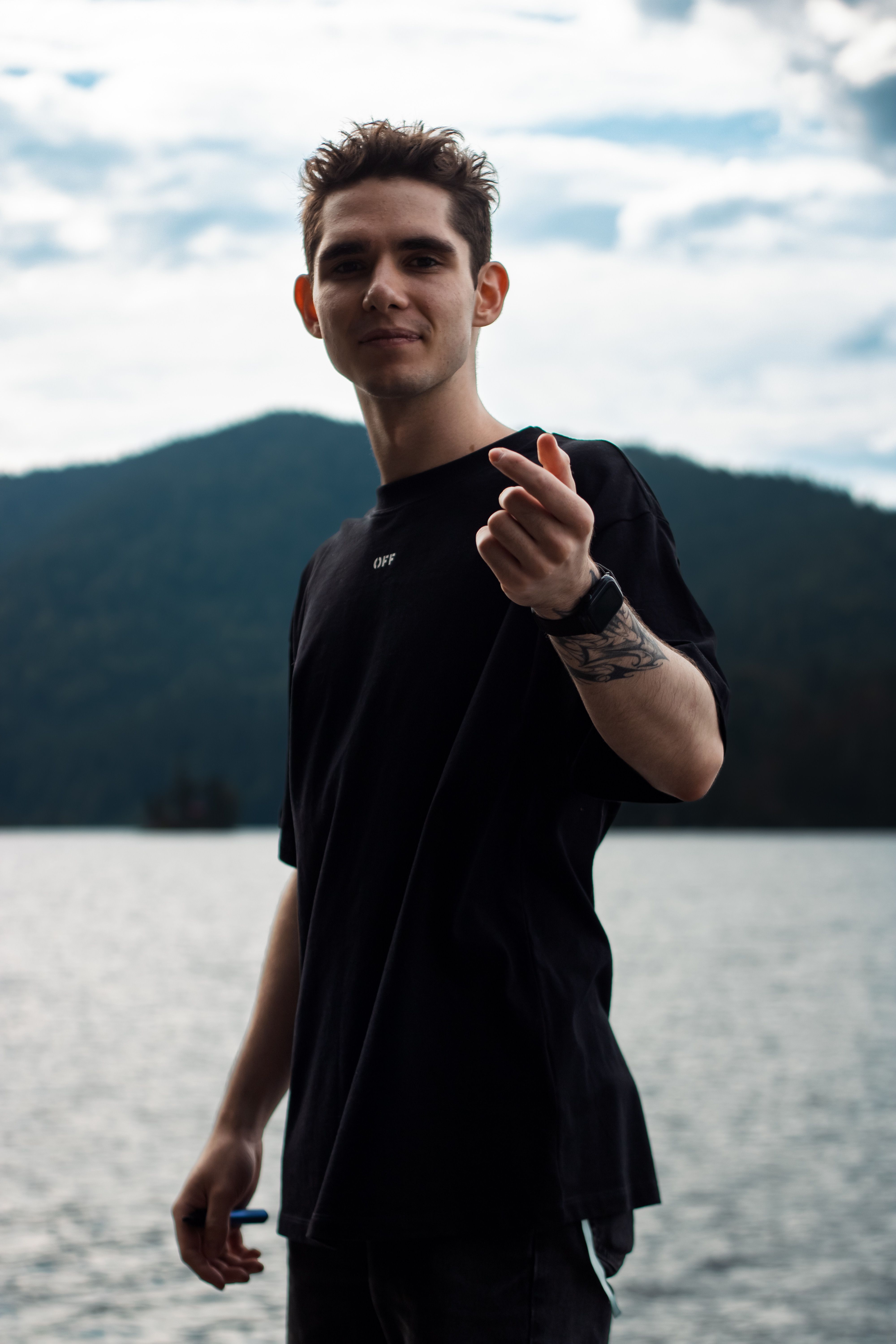 Young man pointing at camera by a serene mountain lake, under a cloudy sky.
