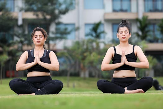 Two women practicing yoga in a peaceful outdoor setting, wearing black athletic wear and sitting cross-legged.
