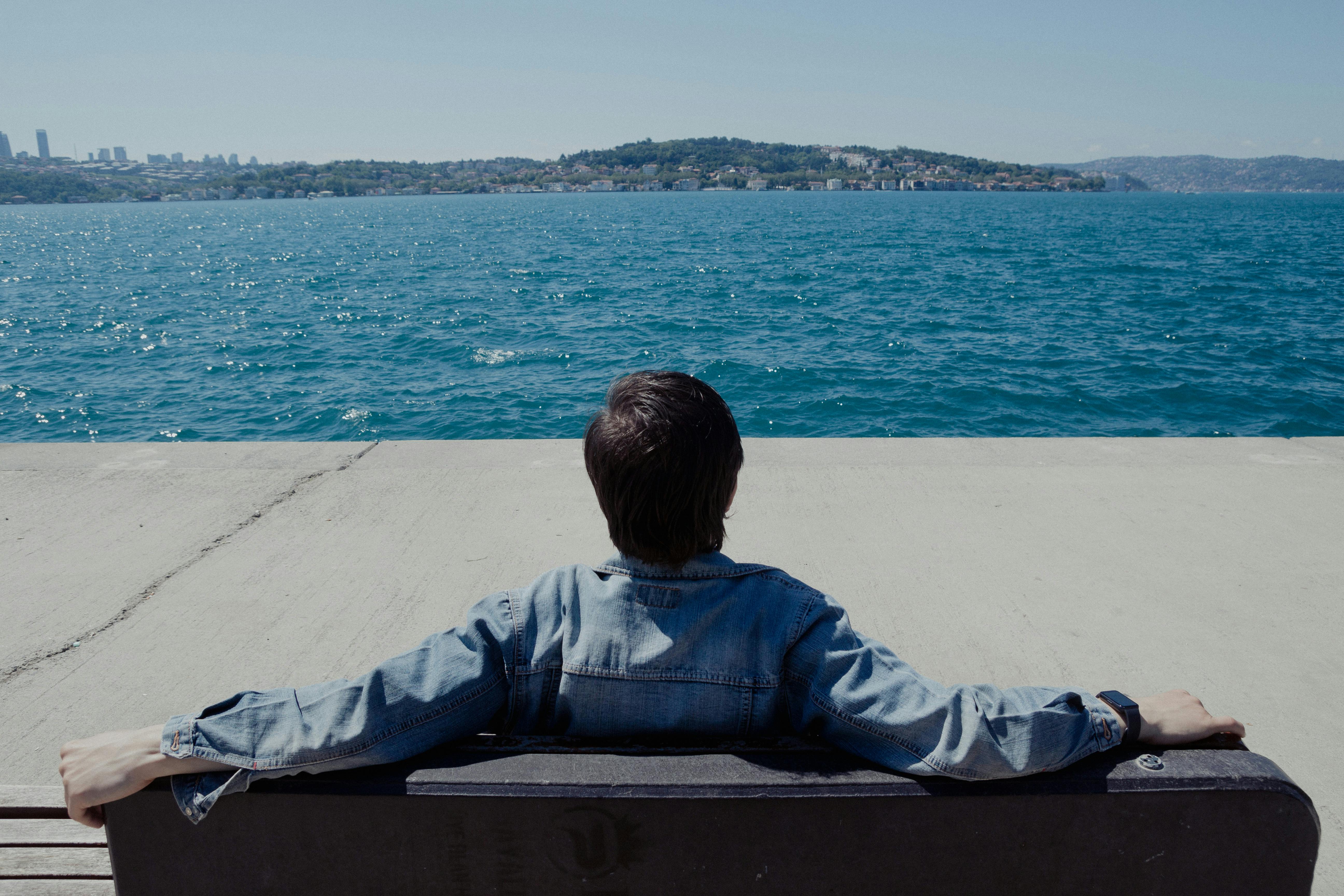 An adult man in denim jacket sits on a bench by the Bosporus, enjoying a sunny day.