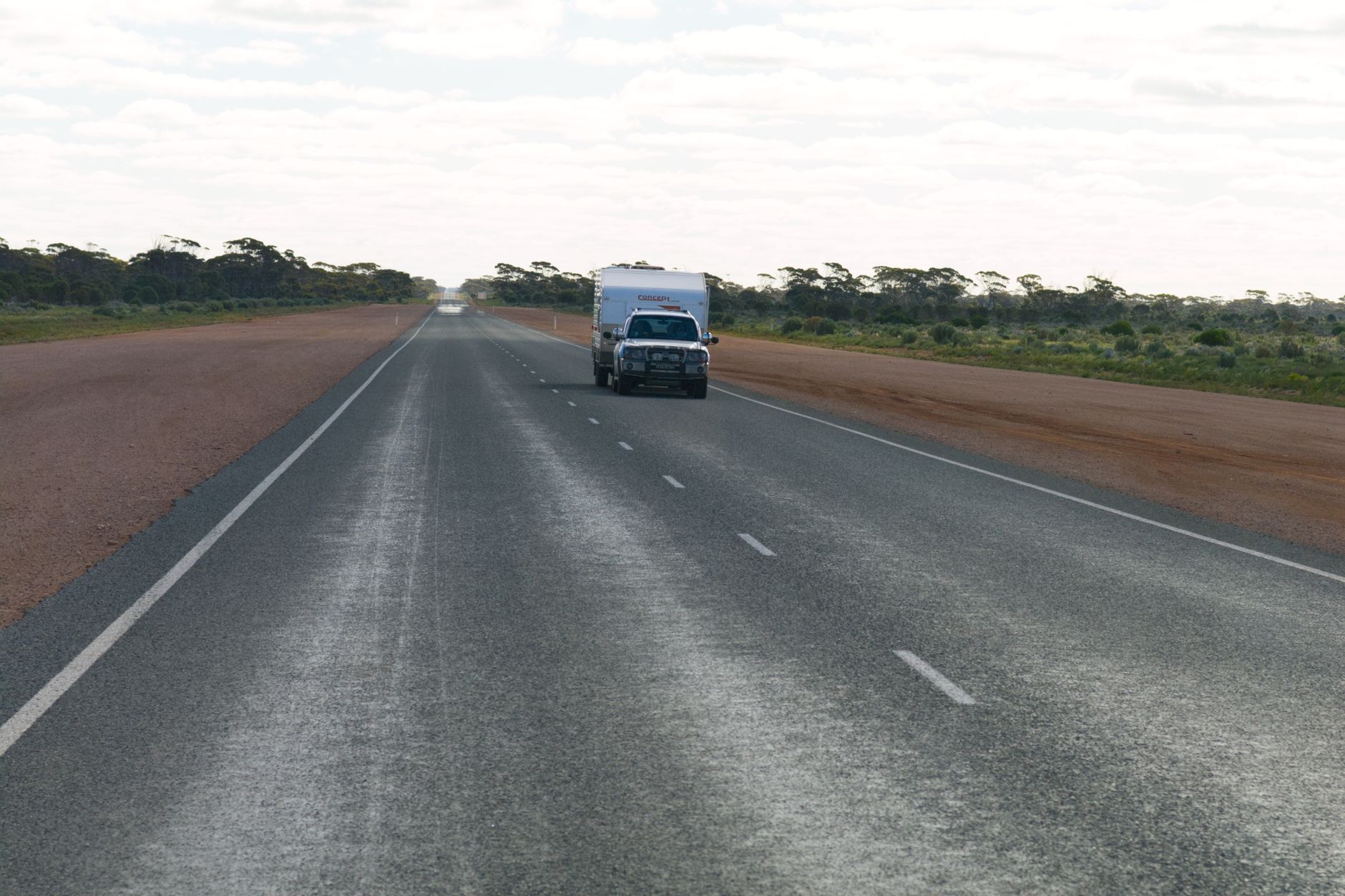 A vehicle travels a deserted road in an expansive rural landscape under a cloudy sky.