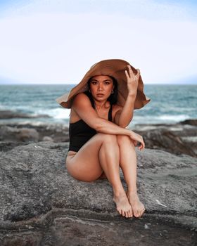 A woman in a sun hat and swimsuit sitting on rocks by the ocean, embodying summer vibes.