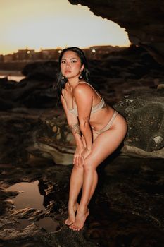 A woman in swimwear poses on rocky beach during a sunset, with a dramatic coastal backdrop.