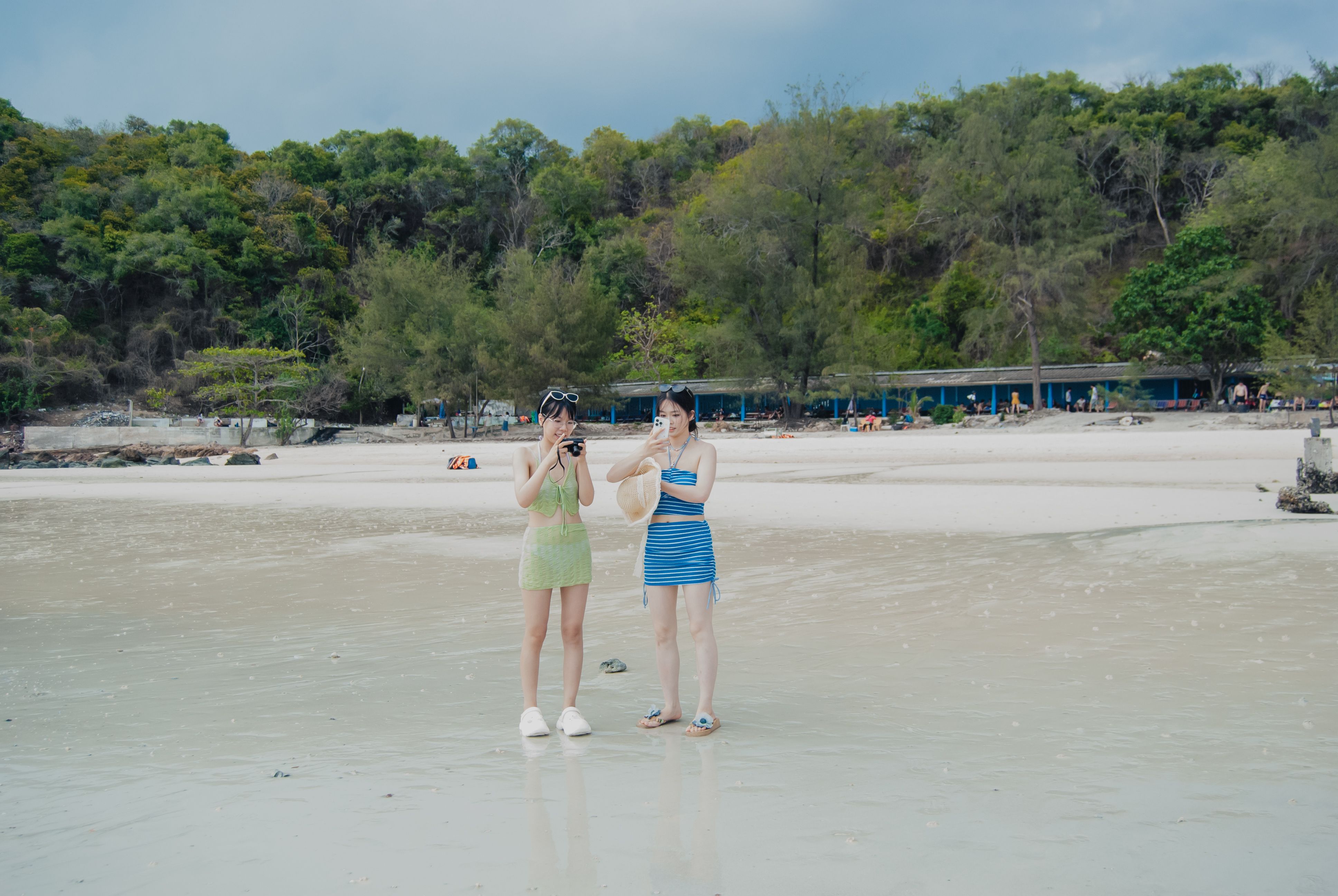 Two women taking photos on a scenic beach with lush green hills in the background.
