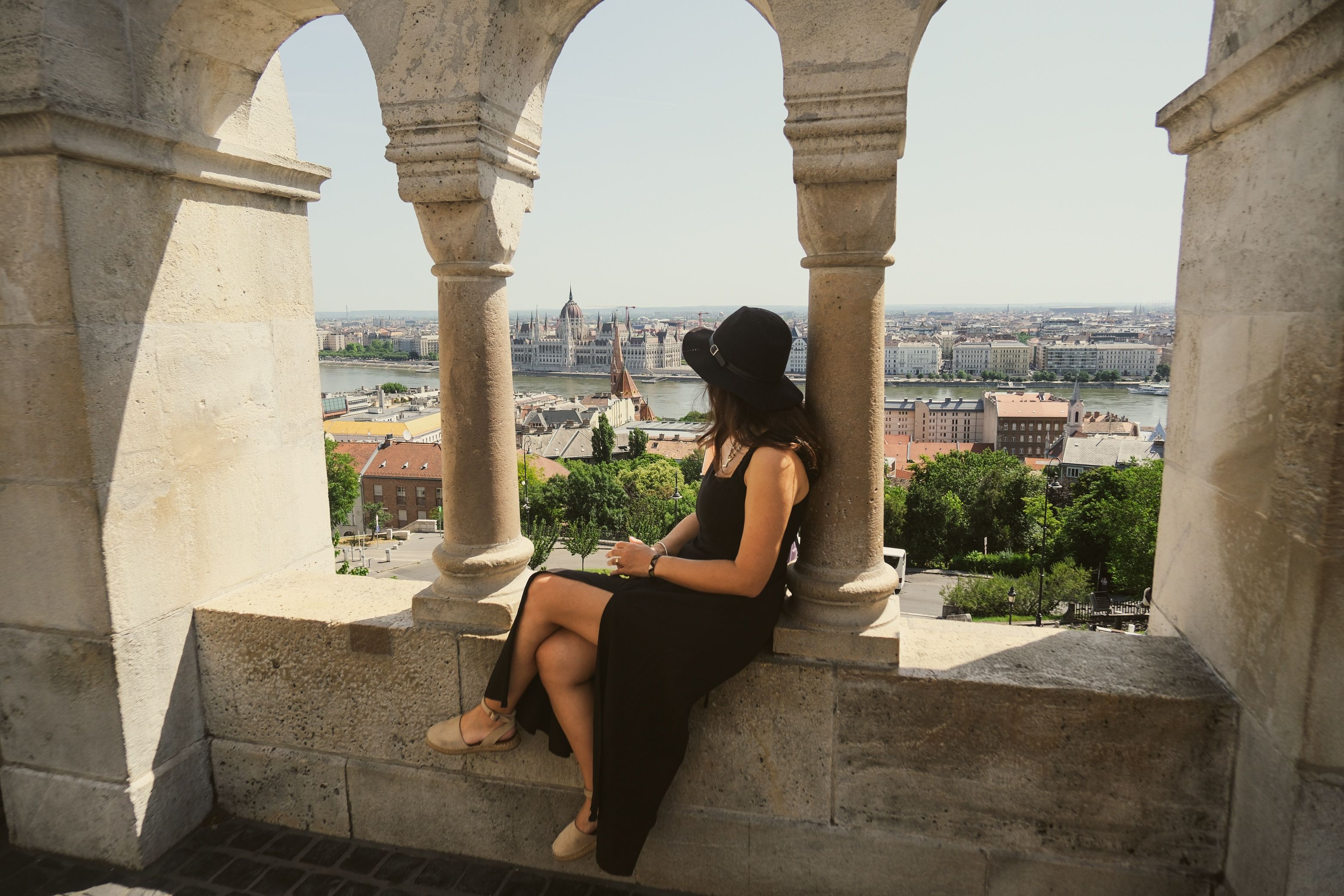 A woman in a black dress enjoys the panoramic view of Budapest from Fisherman's Bastion on a sunny day.