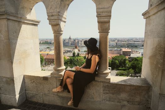 A woman in a black dress enjoys the panoramic view of Budapest from Fisherman's Bastion on a sunny day.