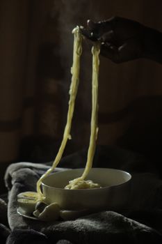 Steam rises from a bowl of freshly cooked noodles being lifted by a hand in a dark setting.