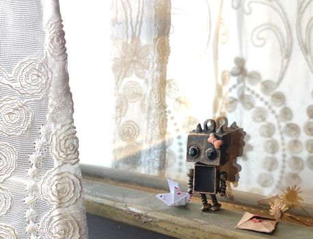 A charming robot figurine with an origami heart sits on a windowsill next to lace curtains, evoking a playful scene.