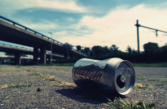 A close-up photo of an empty Coca-Cola can lying on an urban road, symbolizing litter.