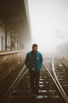 Man in blue jacket walking alone on foggy train tracks at a misty station.
