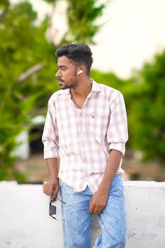 Young man leaning on railing with earbuds, wearing casual attire outdoors.