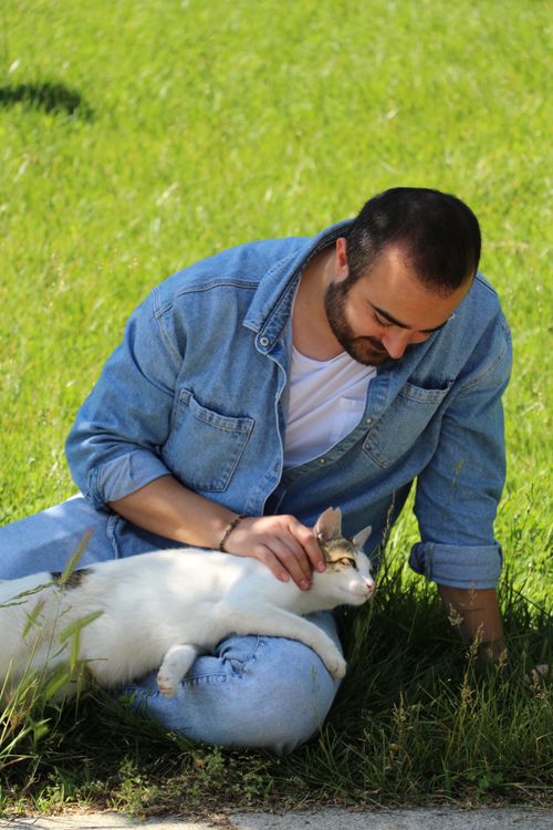 Free A man in denim enjoys a sunny day outdoors while petting a cat on the grass. Stock Photo