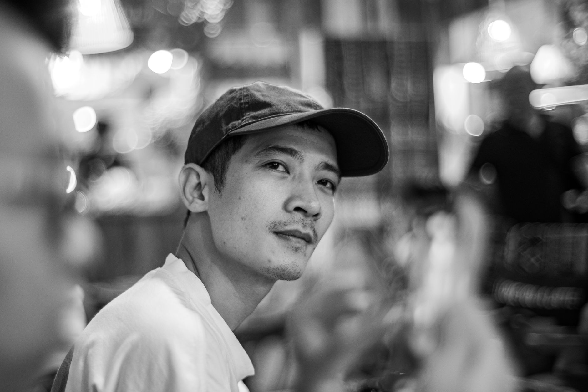 Black and white portrait of a man wearing a cap in a bustling urban cafe setting.