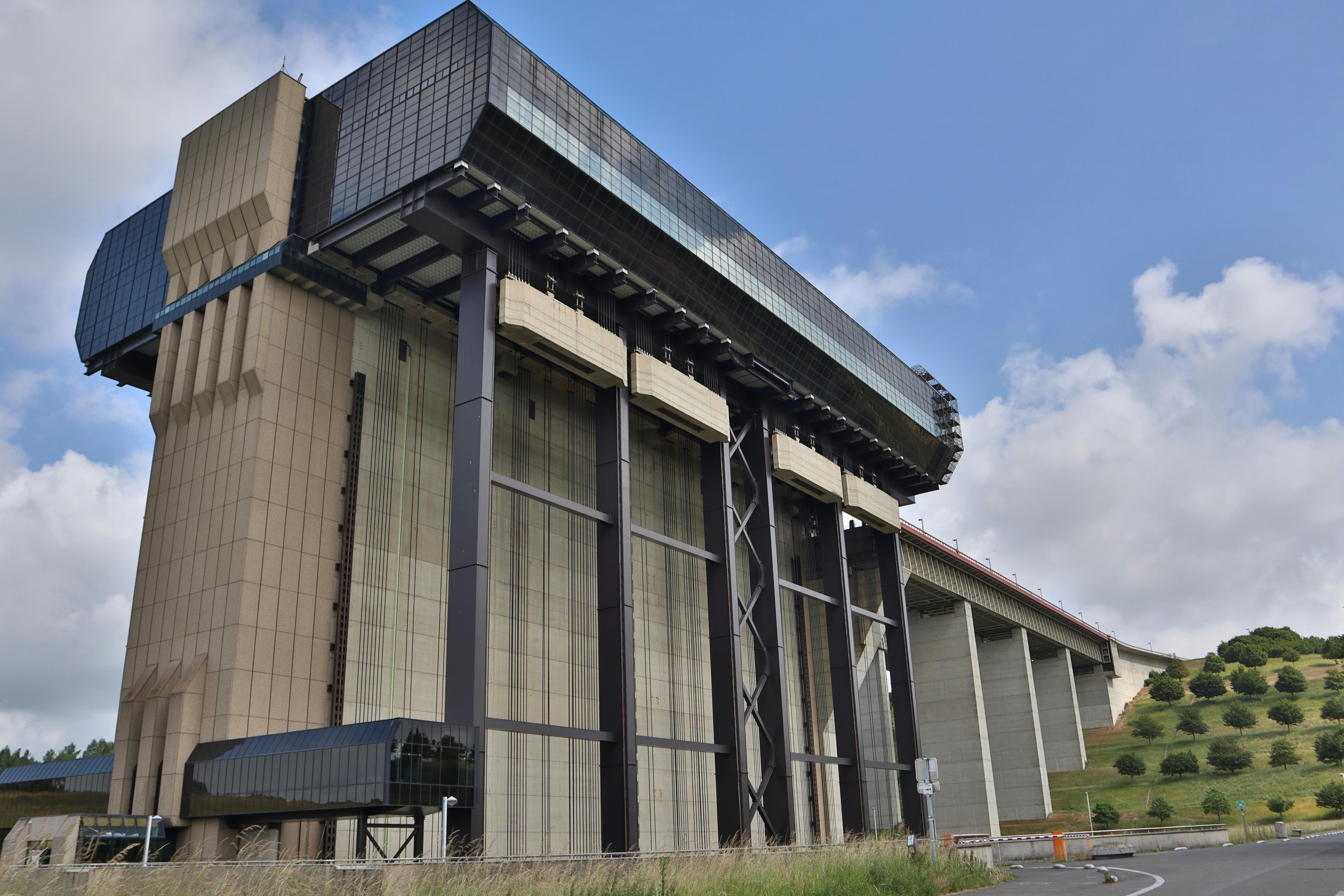 Massive, brutalist, industrial structure of concrete and glass, with an extending bridge, set against green hills and a cloudy blue sky.