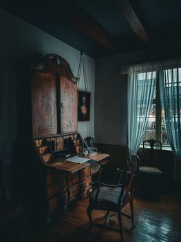Moody vintage study room featuring an antique wooden desk, chair, and window curtains.