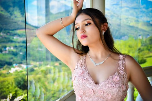 A young woman in a pink floral dress poses elegantly by a window with a scenic view.