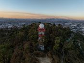 Aerial shot over Monterrey showcasing a radio tower amidst trees with cityscape backdrop at sunset.