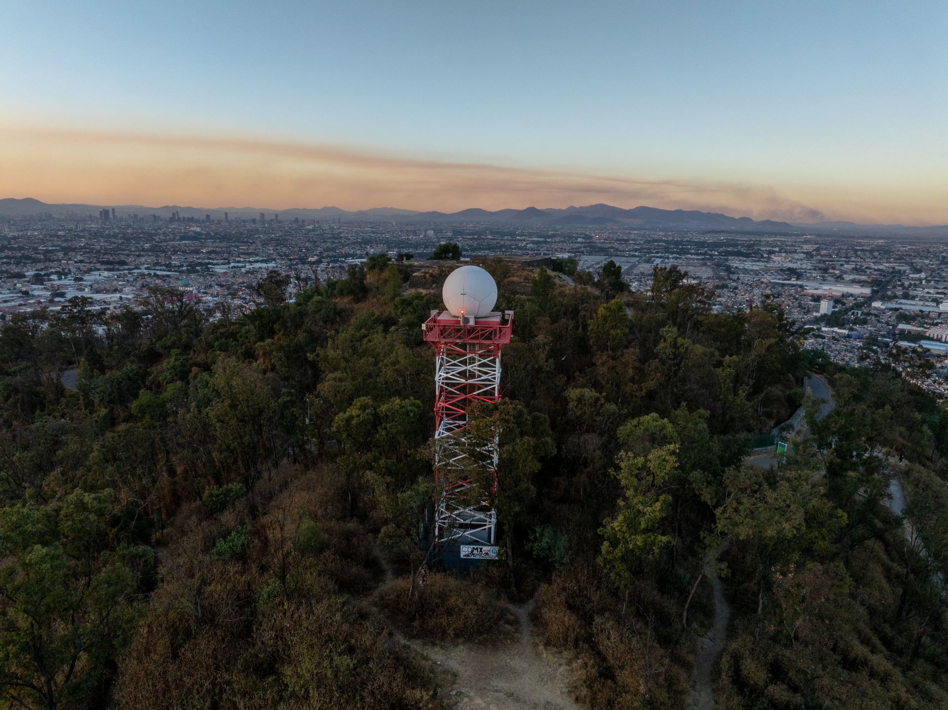 Aerial shot over Monterrey showcasing a radio tower amidst trees with cityscape backdrop at sunset.