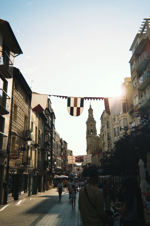Free Bustling street in Spain with historic architecture and flags under evening sun. Stock Photo