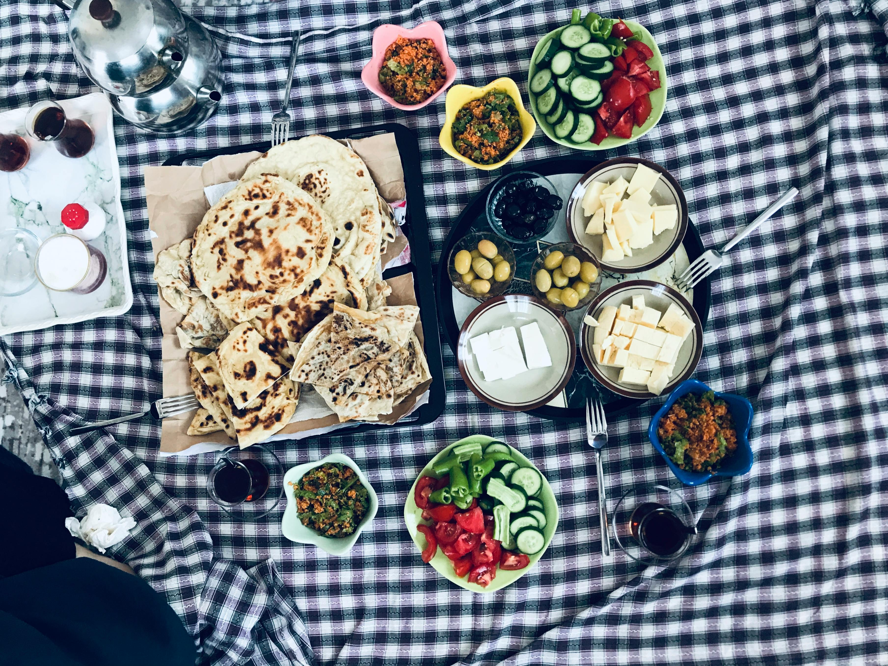 A top view of a vibrant traditional picnic spread featuring cheese, olives, vegetables, and flatbread on a checkered blanket.