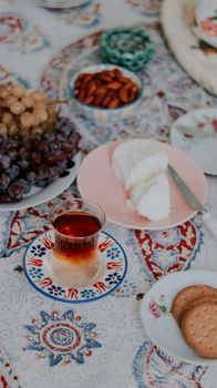 A beautifully arranged table with tea, cheese, grapes, almonds and cookies on a traditional decorative tablecloth.