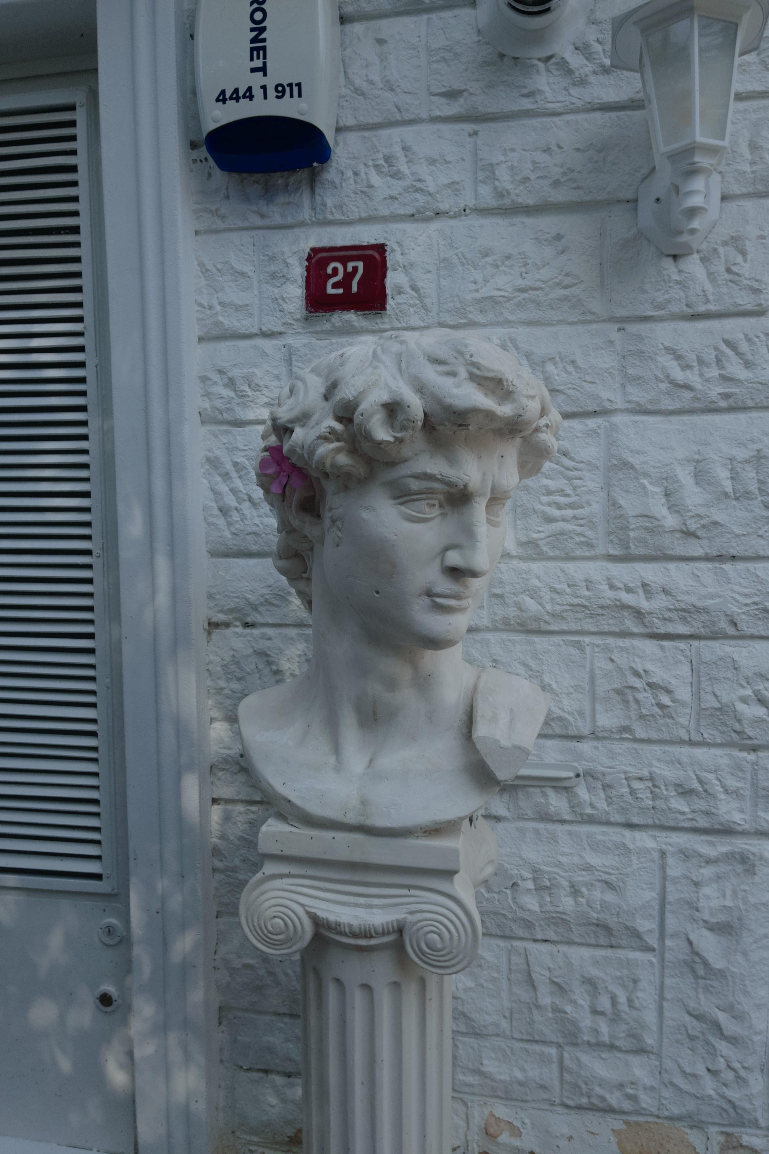 Marble bust of David sculpture against a white brick wall with house number 27.
