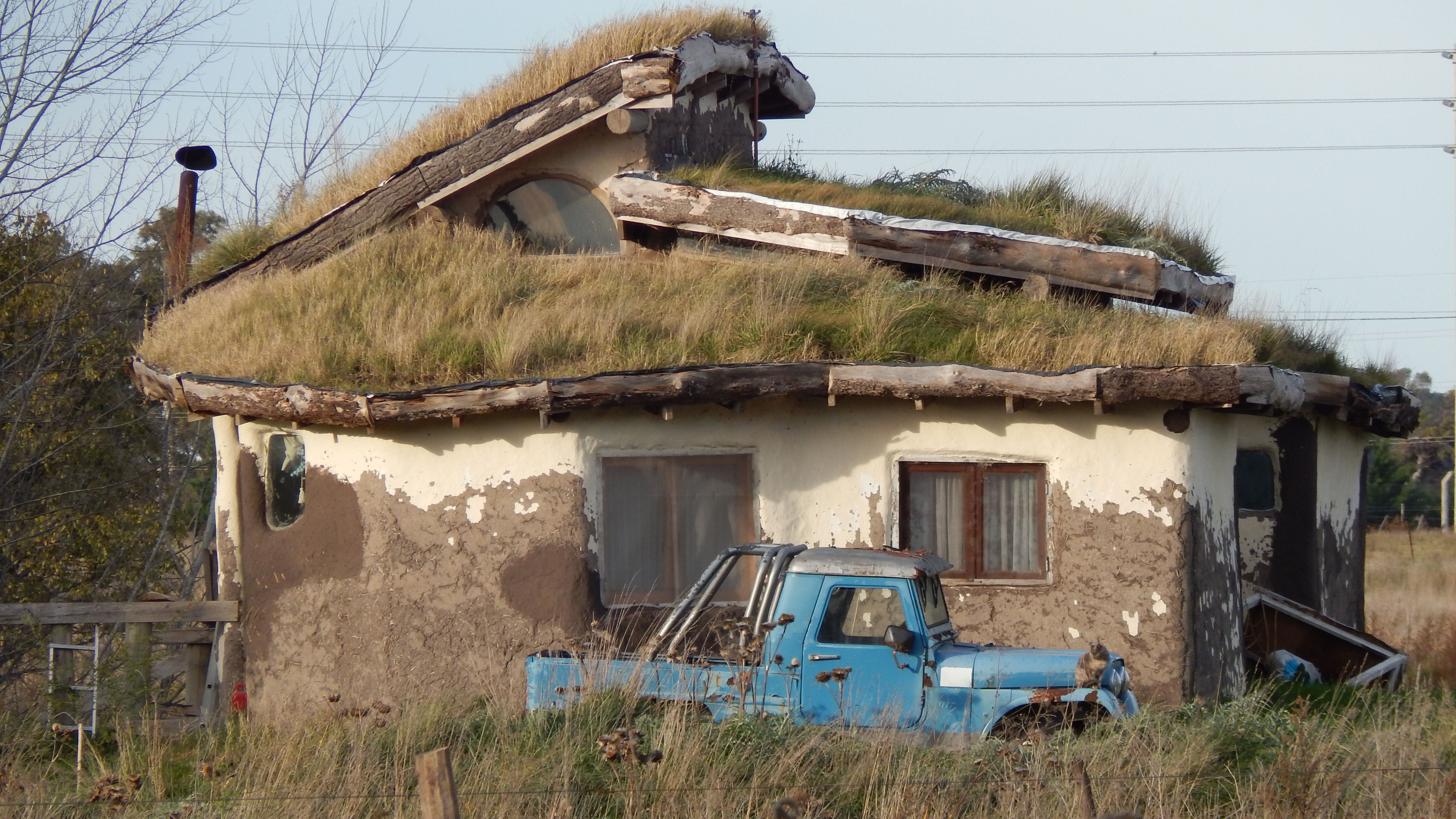 Casa abandonada com telhado coberto de grama e veículo antigo encostado na parede, transmitindo uma sensação de abandono e decadência.