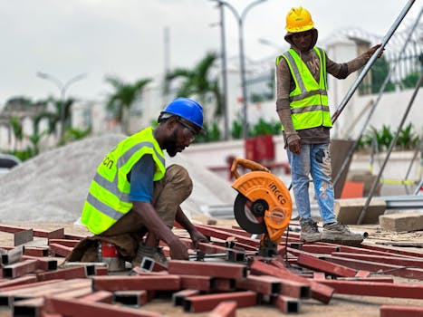 Two male construction workers in safety gear assembling metal framework outdoors.