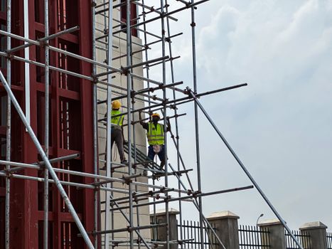 Two workers in safety vests and helmets work on scaffolding, set against a cloudy sky.