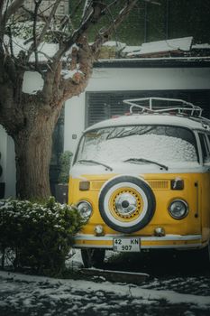 A bright yellow vintage van covered in snow parked under a tree in a residential area.