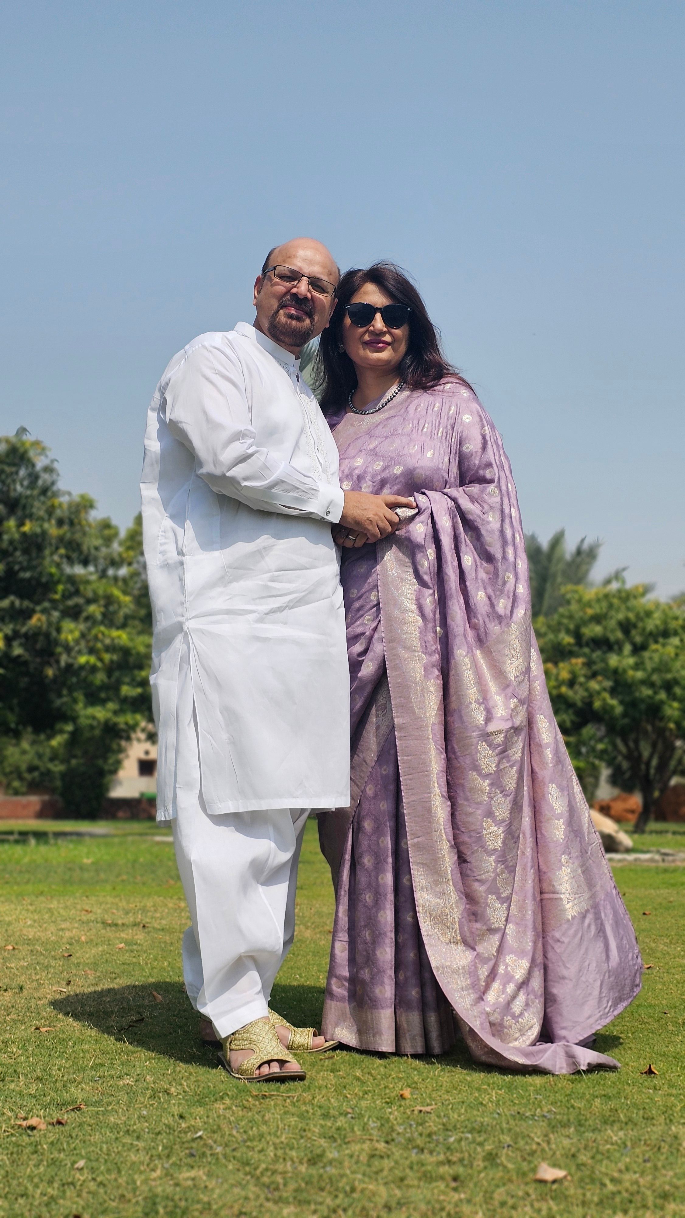 A smiling couple poses in traditional Indian clothing in a sunny outdoor park setting.