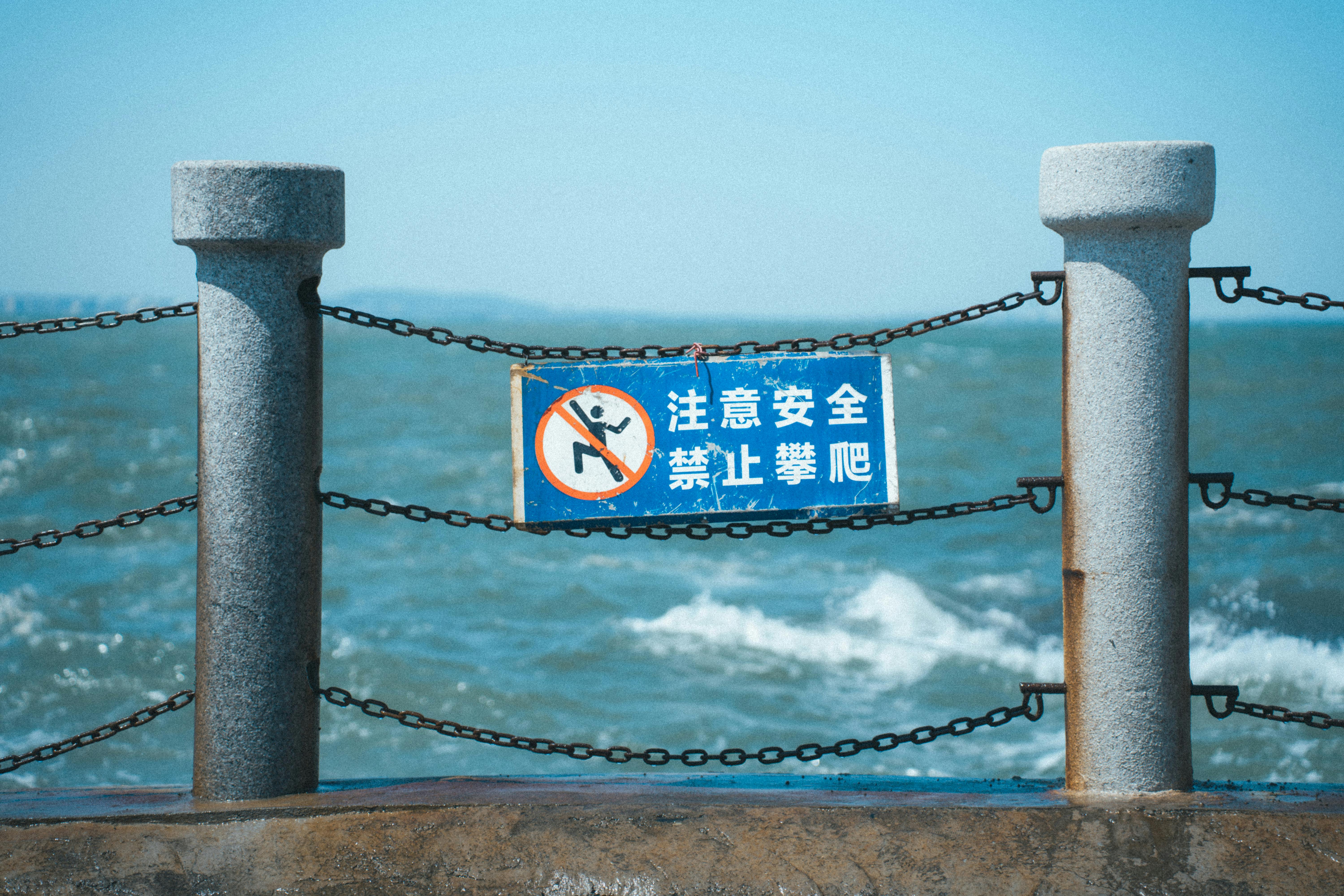 Oceanfront safety sign in Chinese warning against climbing, with turbulent sea waves and blue sky background.