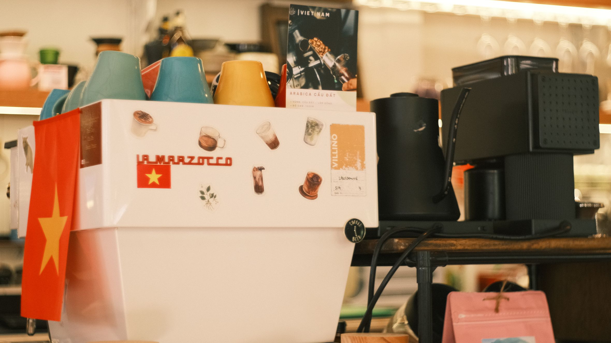A cozy coffee shop with a La Marzocco espresso machine adorned with colorful cups and a Vietnam flag.