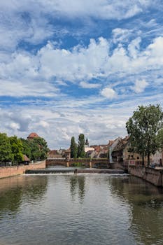 Charming view of a European cityscape with historic architecture, river, and picturesque bridge under a partly cloudy sky.