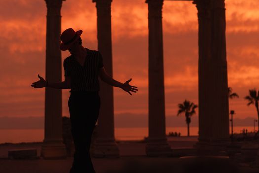 Dramatic silhouette of a performer in a red hat against ancient columns at sunset.