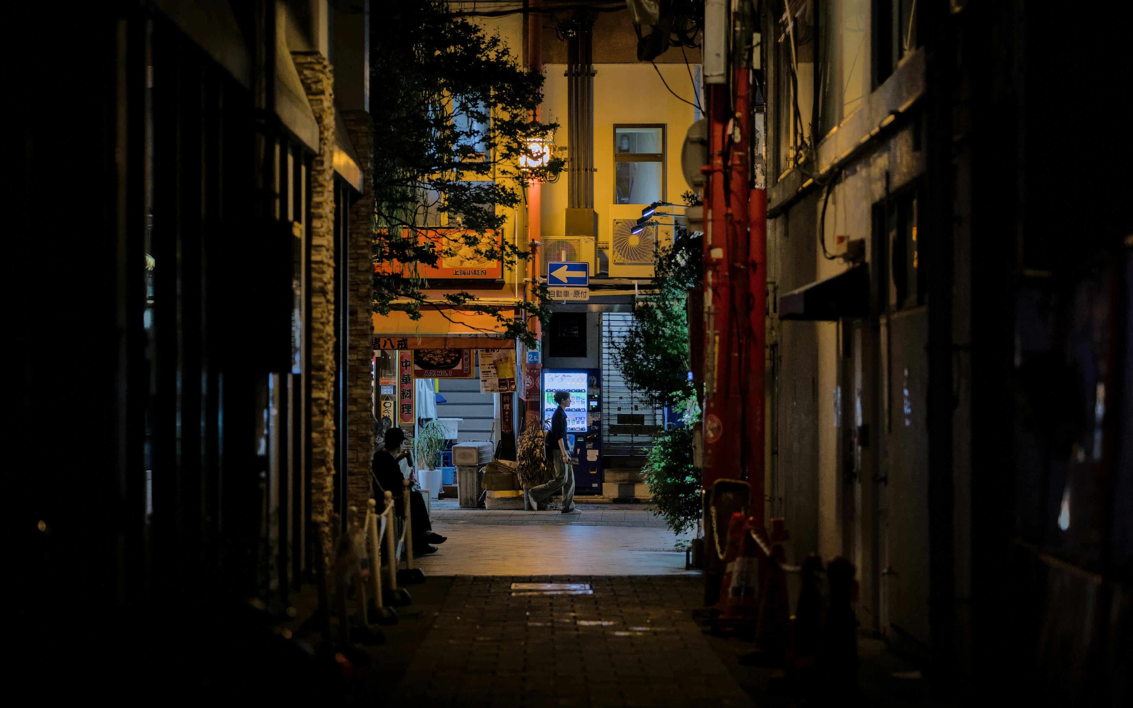 A moody nighttime view of an alley with vending machines and a solitary pedestrian in Kobe, Japan.