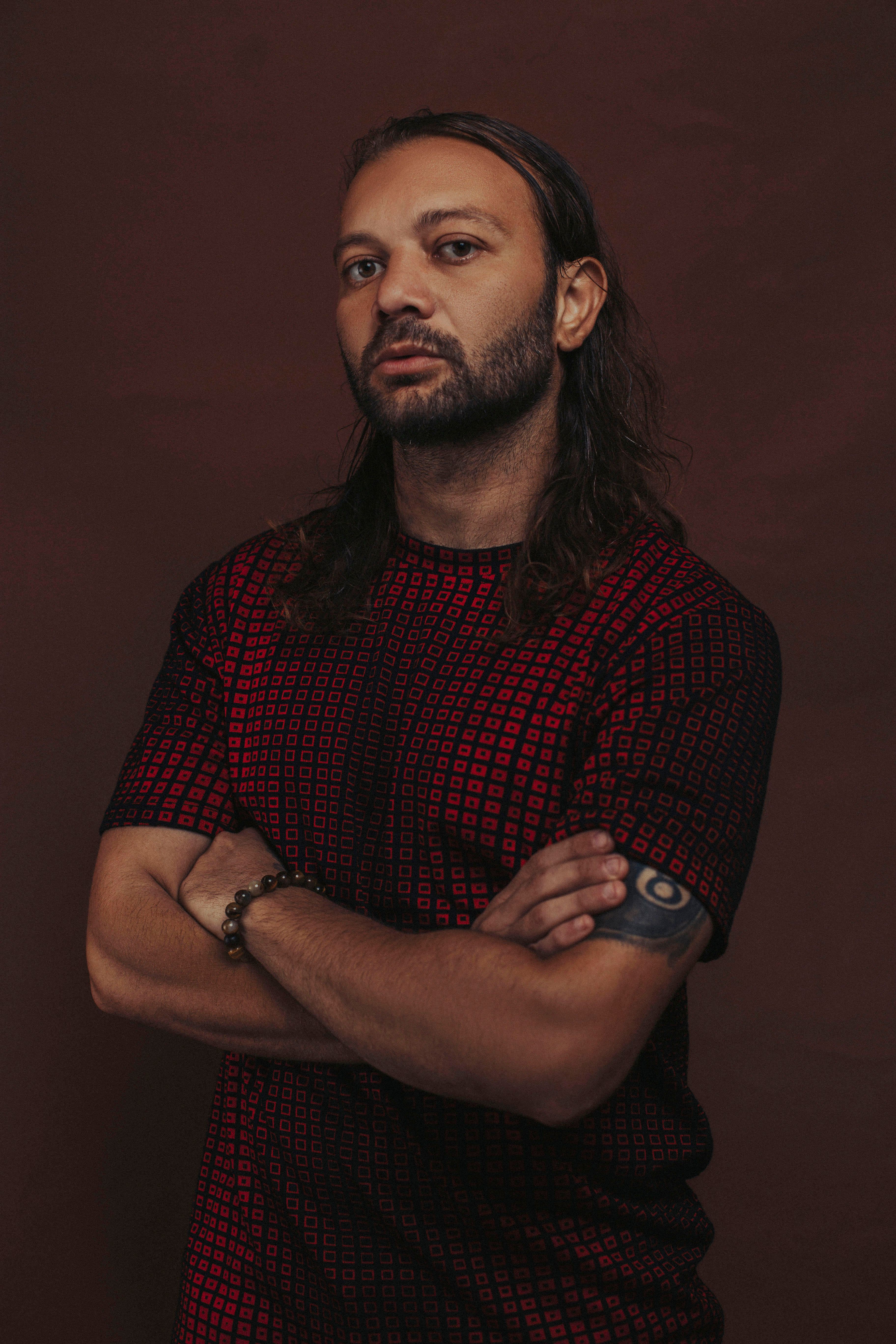 Man with beard and tattoo wearing red geometric shirt, arms crossed.