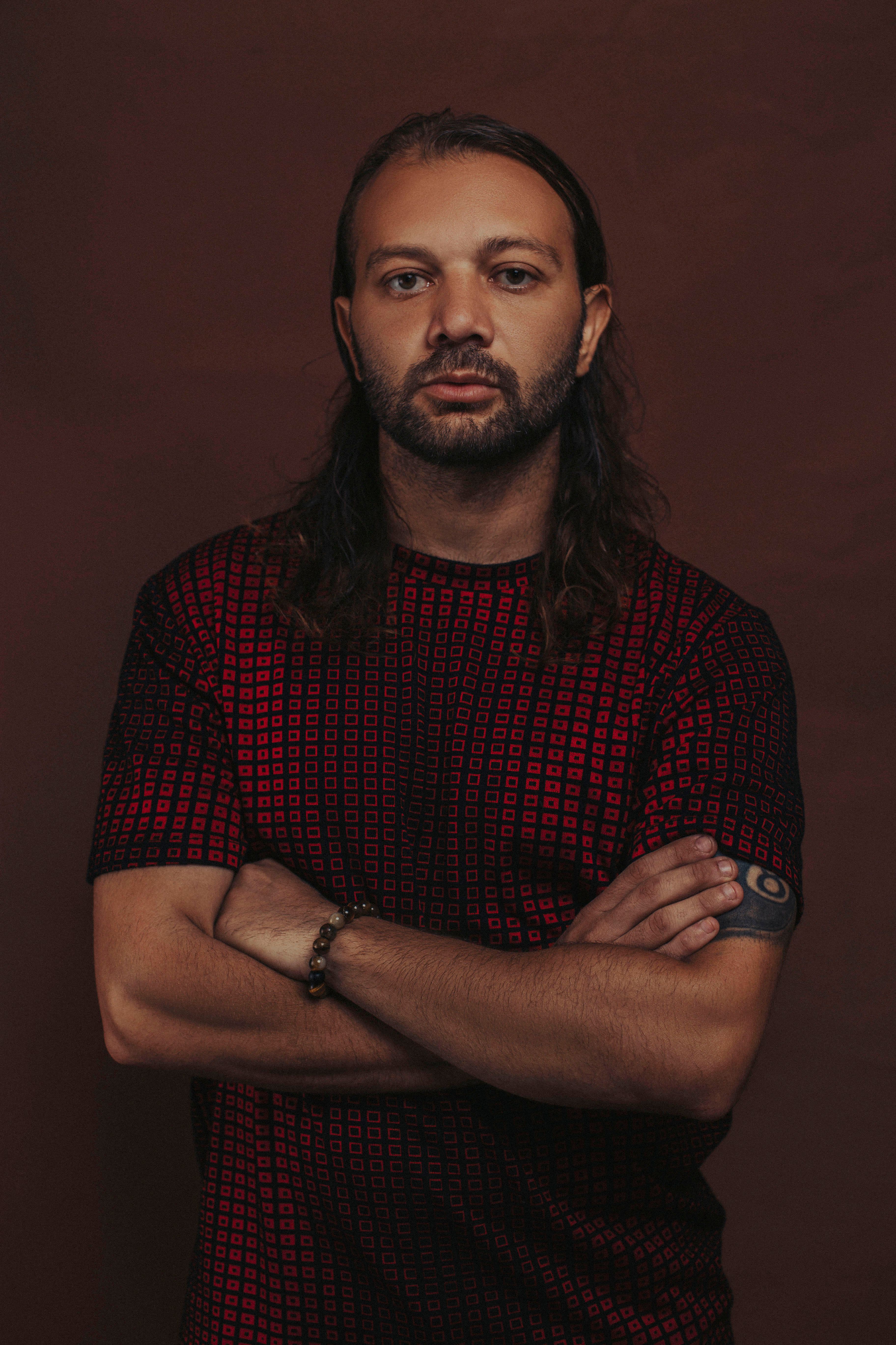 Close-up portrait of a man with long hair and tattoos, arms crossed, wearing a patterned shirt.
