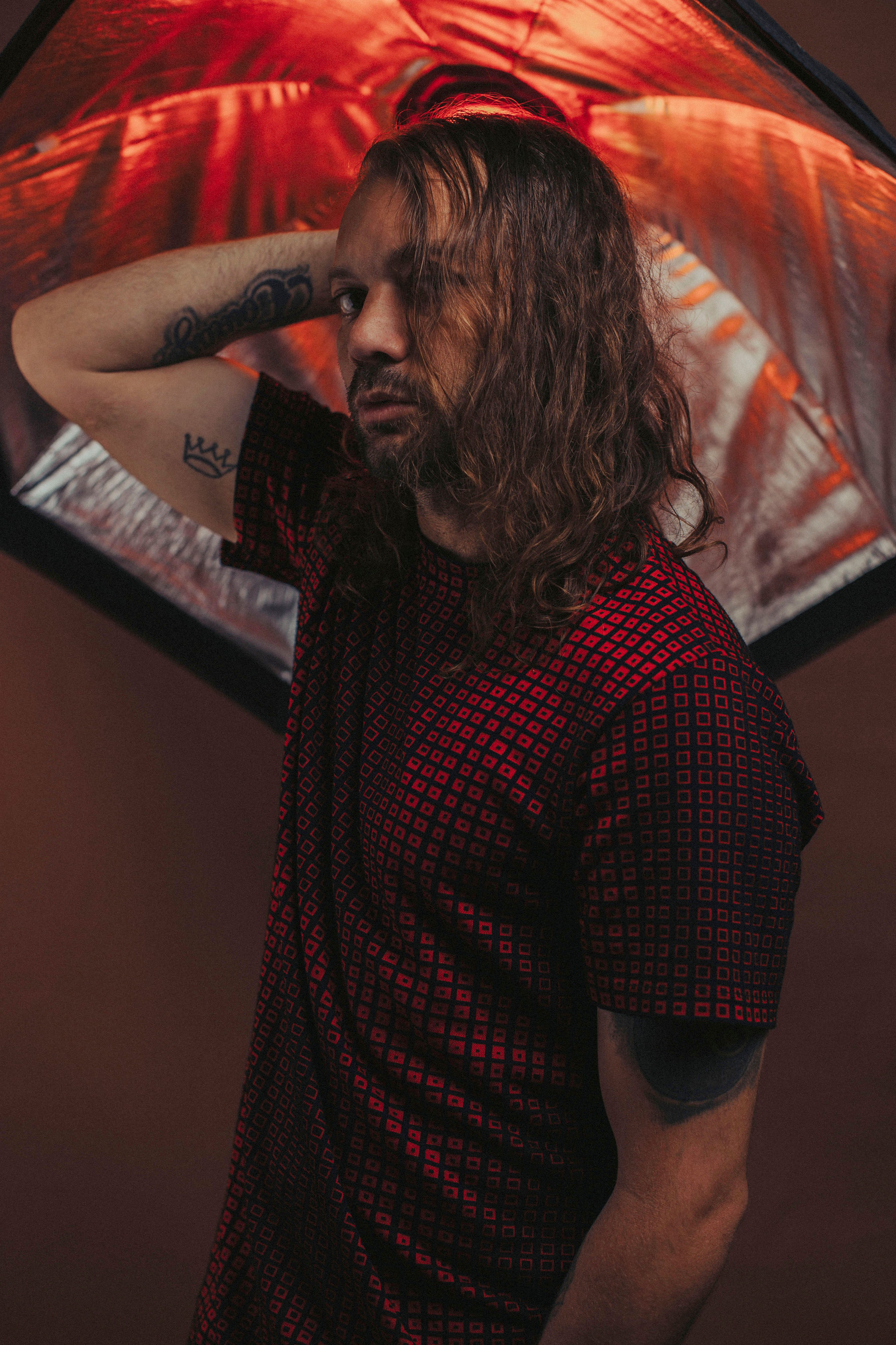 A man in red patterned shirt poses thoughtfully under dramatic lighting.